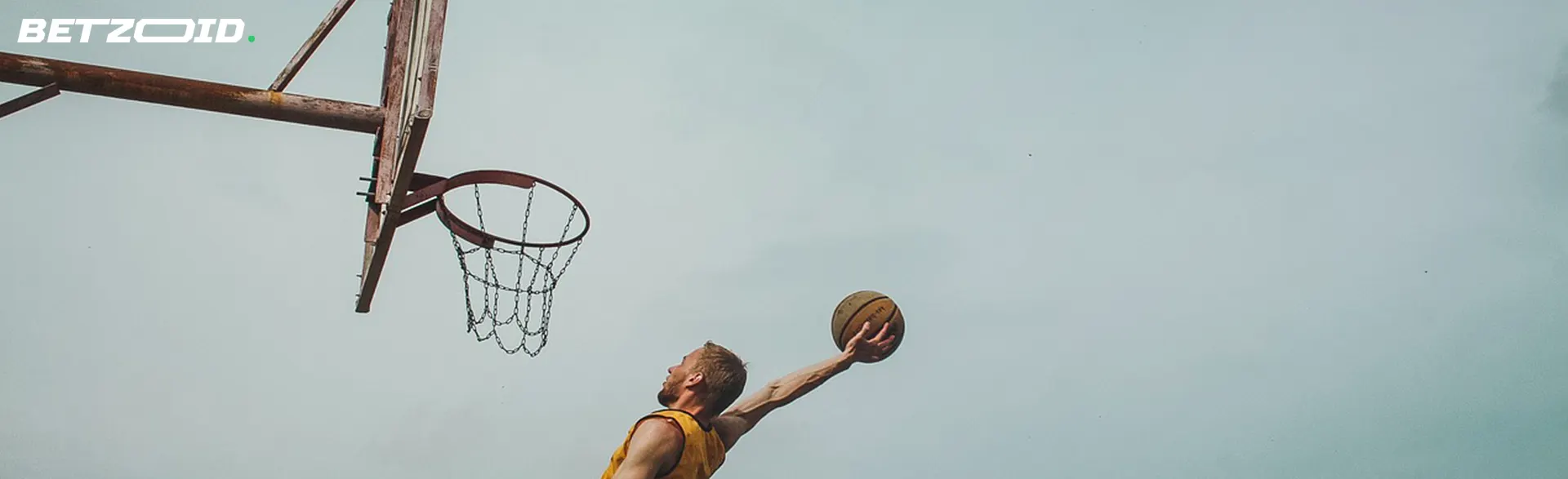 A basketball player in a yellow tank top performing a layup at an outdoor basketball hoop against a clear sky.