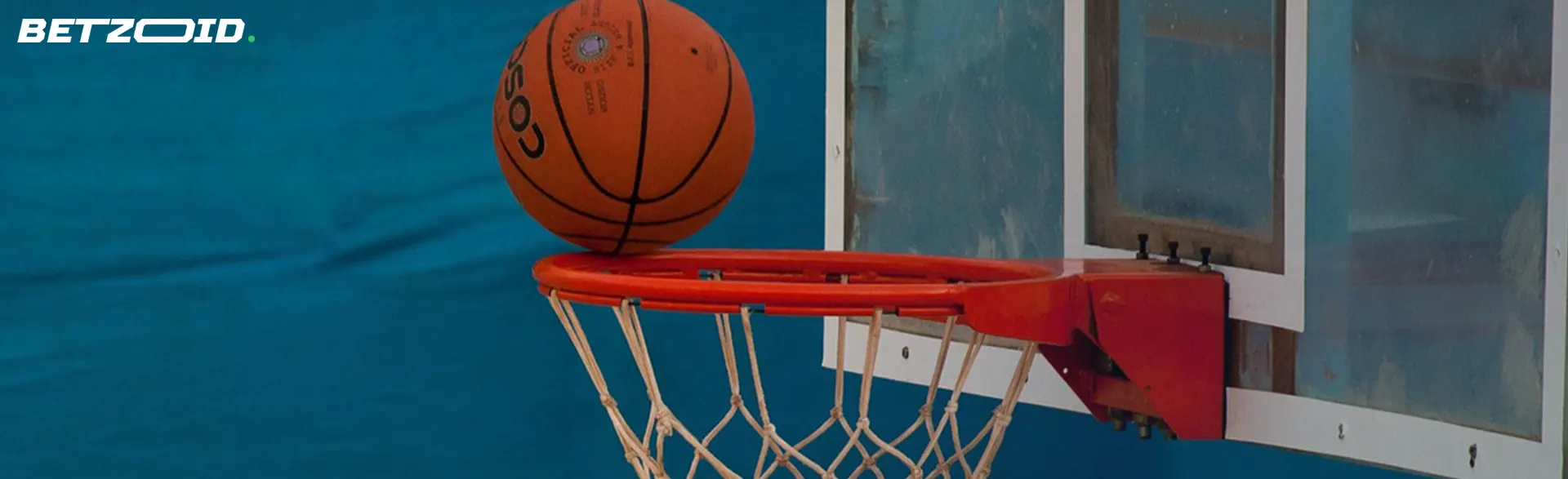 A basketball poised on the rim of a red basketball hoop against a blurred blue background.