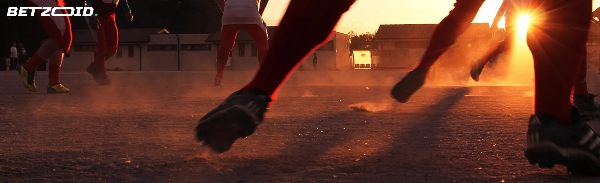 Soccer players in action at sunset, kicking up dust on a dry field.
