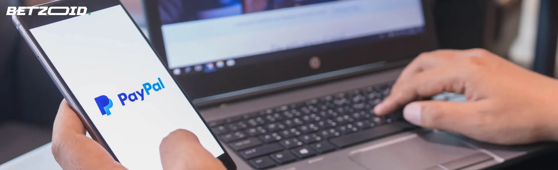 A man uses PayPal on a smartphone in front of a laptop.